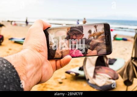 Couple of adult crazy man and woman in video call at the beach doing funny expression - happy people with technology phone device making crazy - joy and nice adults at the beach during vacation Stock Photo