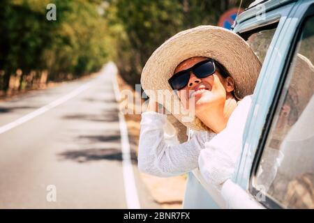 Happy young beautiful caucasian people female enjoy the sun outside the window of blue old vehicle - long road in background for travel and transport concept with happiness and joyful lifestyle Stock Photo