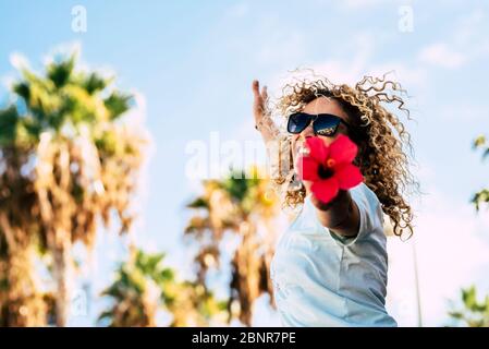 Happiness and red flowers - spring season andhapy people in outdoor leisure activity - blonde curly beautiful people adult woman jumping and showing a big red flowers at the camera Stock Photo