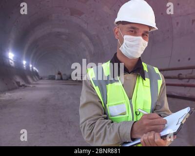 Construction worker dressed in medical mask, uniform and white hardhat stands with clipboard at metro tunnel under construction Stock Photo