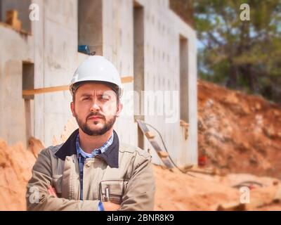 Caucasian bearded civil engineer or construction worker in white hardhat stands cross handed opposite construction site. Stock Photo