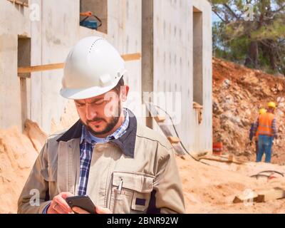 Caucasian bearded civil engineer or construction worker in white hardhat uses his pnone opposite construction site. Stock Photo
