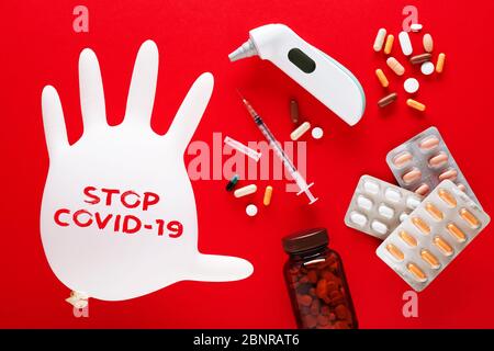 Inflated medical glove with stop covid-19 text message, syringe, pills and thermometer on red background. Corona virus or covid 19 disease treatment o Stock Photo