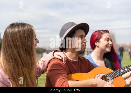 A Hispanic boy in a hat plays the guitar alongside two Caucasian girls in a city park Stock Photo