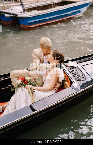 Italy wedding in Venice. The bride and groom ride in a classic wooden gondola along a narrow Venetian canal. Close-up of cuddles newlyweds. Stock Photo
