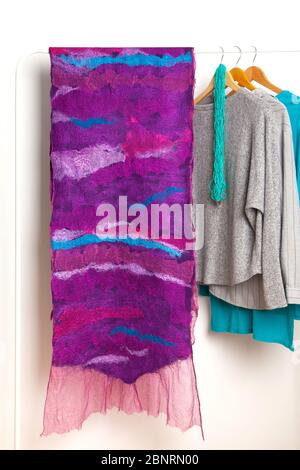 Beautiful large felted purple silk woolen homemade scarf hanging on white hanger next to gray blue shirts on white background in studio. Spring wardro Stock Photo