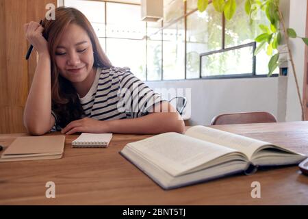 Closeup image of a woman holding a pen to write on a notebook and textbooks on wooden table while studying Stock Photo