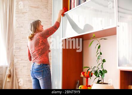 Side view of adult lady in casual clothes removing dust with cloth from shelf while cleaning cozy room at home Stock Photo