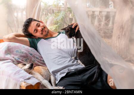 young man reclining on an armchair in the garden behind a mosquito net, strokes a black cat sitting on his side Stock Photo