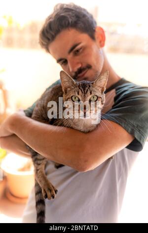 vertical photo. Young man  hugs a green-eyed tabby cat. The cat looks at the camera. Stock Photo