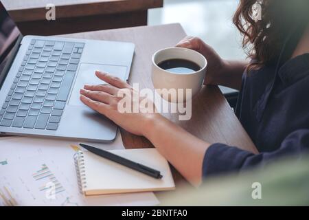 Closeup image of a woman touching on laptop touchpad while drinking coffee and working in office Stock Photo