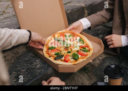 Close-up of Couple grab slices of pizza from box at the outdoor. Man and woman hands taking pizza. Vegan pizza with fresh tomatoes basil and broccoli Stock Photo