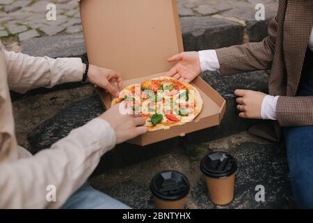 Close-up of Couple grab slices of pizza from box at the outdoor. Man and woman hands taking pizza. Vegan pizza with fresh tomatoes basil and broccoli Stock Photo