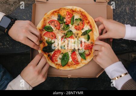 Top view image of couple grab slices of pizza from box at the outdoor. Man and woman hands taking pizza. Vegan pizza with fresh tomatoes basil and Stock Photo