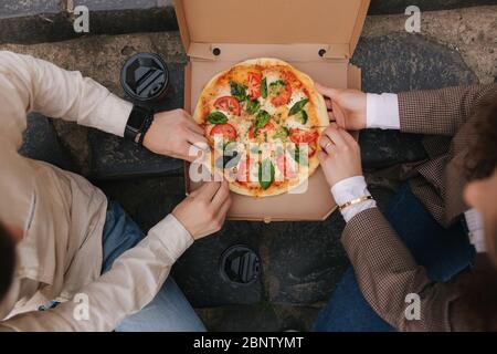 Top view image of couple grab slices of pizza from box at the outdoor. Man and woman hands taking pizza. Vegan pizza with fresh tomatoes basil and Stock Photo