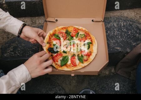Man grab slices of pizza from box at the outdoor. Male hands taking pizza. Vegan pizza with fresh tomatoes basil and broccoli. Lactose and gluten free Stock Photo