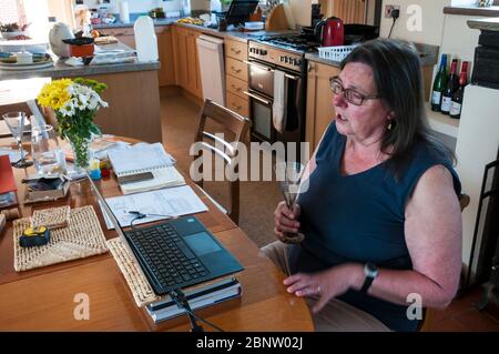 A middle-aged Caucasian woman chatting to her friends on a Zoom conference call or cloud meeting from a laptop in her kitchen during the 2020 COVID-19 coronavirus pandemic lockdown. Stock Photo