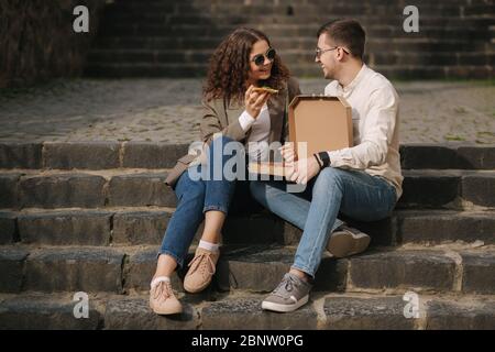 Young couple sitting on stairs outdoor and eating pizza. Handsome man with beaufitul woman taste pizza with vegetables. Vegan fast food