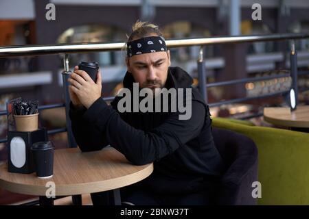 Young handsome lonely man sits cafe table sad waiting for meeting drinking coffee from a paper cup. Caucasian stylish guy dressed black hoodie bandana Stock Photo