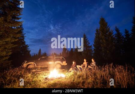Group of friends near bonfire, pickup offroad truck at night camp in the mountains surrounded by spruce trees under magical starry sky. Car travel and night camping concept. Stock Photo