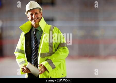 Portrait of smiling mature architect wearing a hardhat and reflective clothing holding a digital tablet on construction site Stock Photo