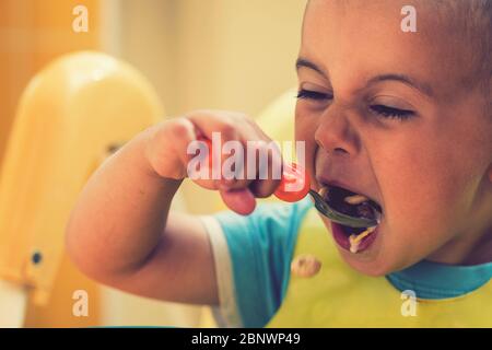 The boy 2 years eats porridge. Children's table. The concept of the child's independence. funny kid in a baby seat. toned Stock Photo