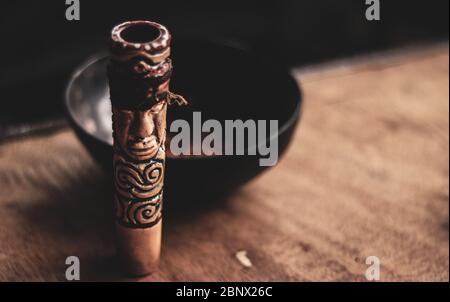 Close up of a brown, vintage Indian pot smoking pipe (chillum) with an engraved tribal design, standing upright on a wooden table with a blurred crush Stock Photo