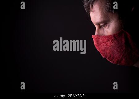 Part of a face en profile of a caucasian male looking down with a homemade wrinkled red cloth face mask. Low key studio COVID-19 outbreak lifestyle Stock Photo