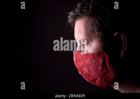 Profile of a caucasian male looking down with a homemade wrinkled red cloth face mask. Low key studio COVID-19 outbreak lifestyle concept. Stock Photo
