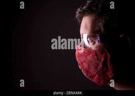 Profile of a caucasian male looking straight forward through reflecting protective plastic glasses and a homemade wrinkled red cloth face mask on. Stock Photo