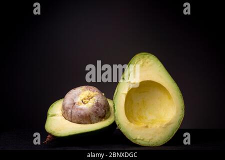Two halves of a large avocado fruit with a huge seed and ripe vibrant yellow green pulp. Studio low key food still life against a dark grey background Stock Photo