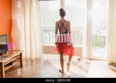 Graceful ballerina girl wearing black leotard, red chiffon midi skirt and pointe ballet shoes, stand face to window with morning soft sun light, train Stock Photo