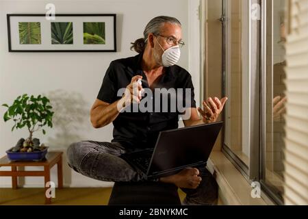 coronavirus working at home concept, man wearing medical mask spraying hand sanitizer using a laptop working from home during the covid 19 pandemic Stock Photo
