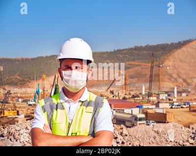 Builder in medical masl and hardhat stands cross handed opposite construction site. Stock Photo