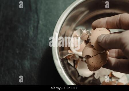 A male hand dips eggshells in a metal bowl. A metal bowl with eggshell on a black background. Adult man is cooking. View from above. Focus in the fore Stock Photo