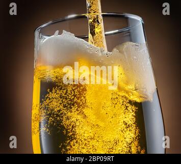 Jet of beer is poured into a beer glass, causing a lot of bubbles and foam. Dark brown background. Stock Photo