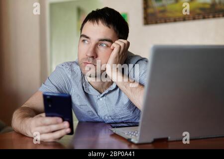 Sad cute young man holding a smartphone in his hand a man working on a laptop remotely. Fatigue from quarantine, self-isolation. Stock Photo