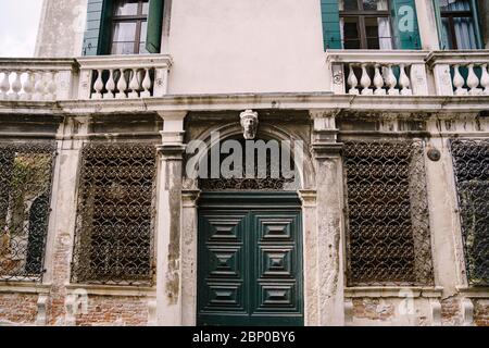Closeup of the facade of a building, on the streets of Venice, Italy. Antique wooden green door. Forged patterns on a metal grate. Balconies with Stock Photo