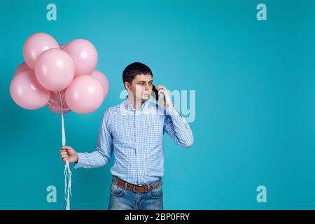 handsome caucasian man in shirt with pastel pink air balloons using smart phone on blue background. copy space Stock Photo
