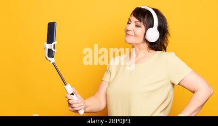 Beautiful Senior woman making video call by smartphone, monopod and wireless headphones over yellow background. Stock Photo
