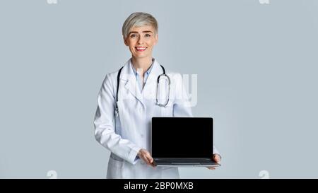Smiling female doctor in white coat shows a laptop with a blank screen. Stock Photo