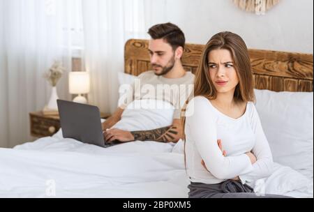 Man works on laptop, woman is offended by bed in bedroom. Stock Photo