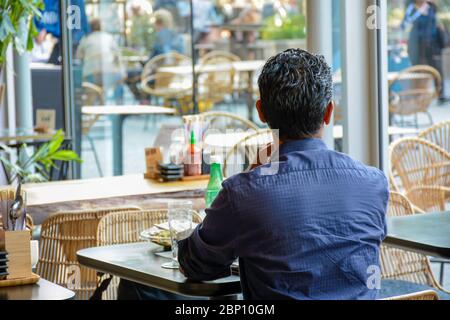 Man from behind sits alone in a modern cafe restaurant and looks outside. He is waiting for his companion to return. There is no one else around him. Stock Photo