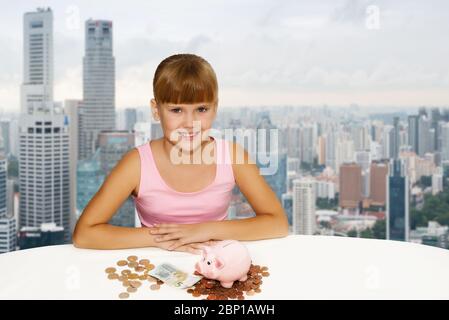Little lovely girl with coinsand piggy bank on the desk over city background. Saving Money for Education Fund. People, children, business concept Stock Photo