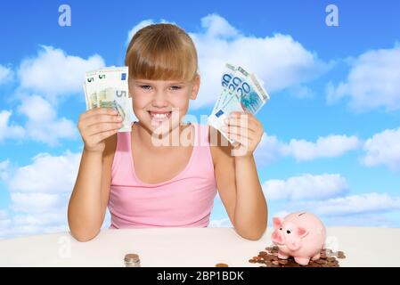 Little cheerful girl sitting on the desk with coins and pink piggy bank and holding in hands european currency over blue sky background Stock Photo