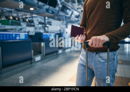 Passenger against check-in counter at airport. Hands of young man holding suitcase and passport. Stock Photo