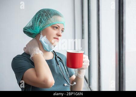 Young caucasian female doctor nurse in surgical mask looks tired. Coffee break during coronavirus covid-19 outbreak Stock Photo