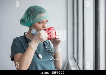 Young caucasian female doctor nurse in surgical mask looks tired. Coffee break during coronavirus covid-19 outbreak Stock Photo