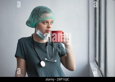 Young caucasian female doctor nurse in surgical mask looks tired. Coffee break during coronavirus covid-19 outbreak Stock Photo