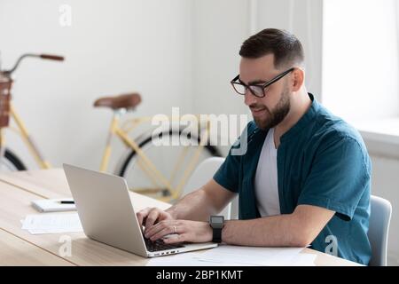 Happy young confident bearded businessman typing on keyboard using laptop. Stock Photo